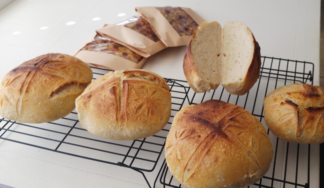 Flower-scored loaves on a rack, one cut open to show the crumb, with bagged focaccia behind